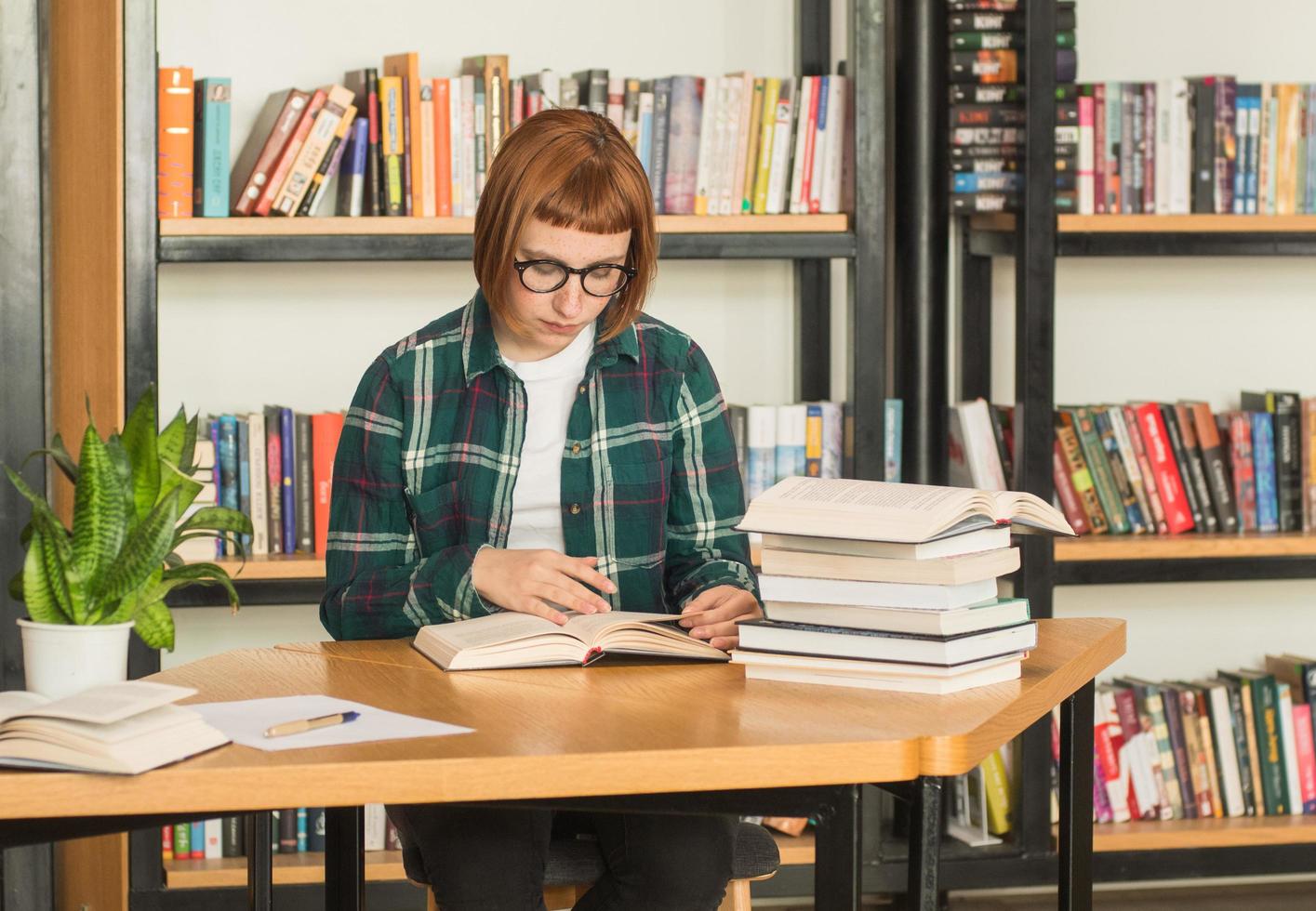 giovane donna rossa con gli occhiali legge il libro in biblioteca foto