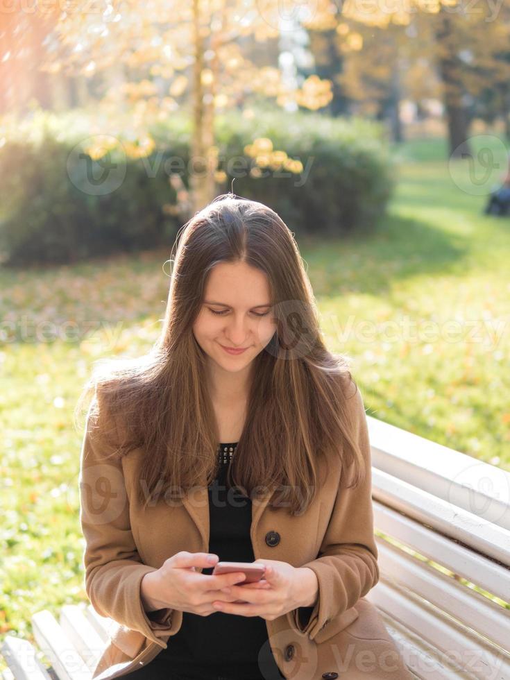 bella ragazza adolescente seduta nel parco su una panchina, in possesso di uno smartphone e in chat online su internet. donna con i capelli lunghi foto