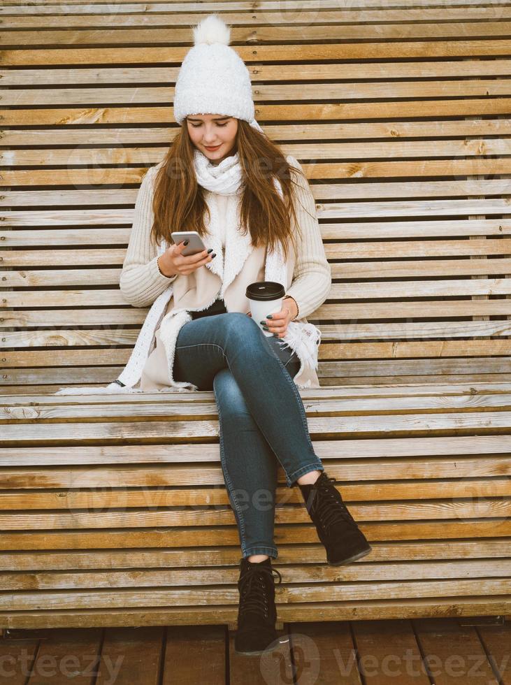 bella ragazza che beve caffè, tè dalla tazza di plastica in autunno, inverno e parla al cellulare. donna con i capelli lunghi seduta su una panchina in autunno o in inverno, crogiolandosi in una bevanda calda, copia spazio foto