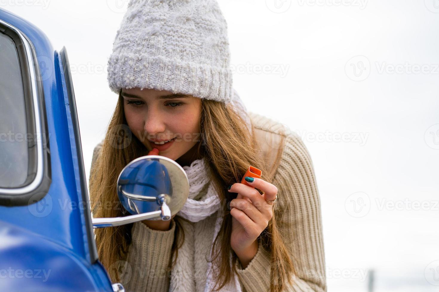 bella ragazza guarda nello specchietto retrovisore dell'auto e trucca le labbra. donna con i capelli lunghi in cappello e sciarpa accanto all'auto retrò blu in autunno o inverno, copia spazio foto