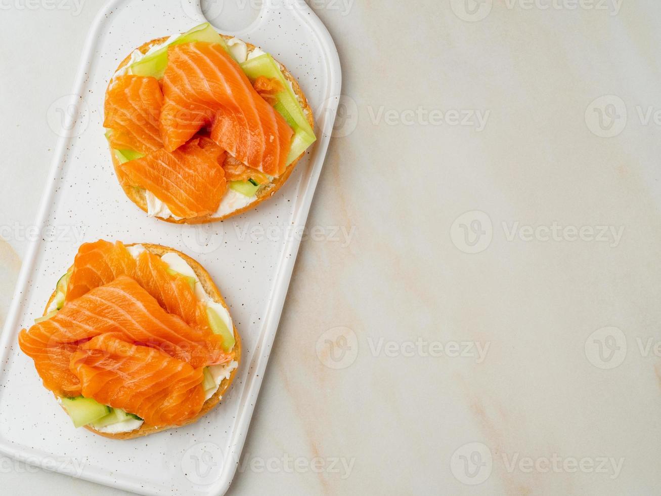 due panini aperti con salmone, crema di formaggio, fette di cetriolo su tavola di marmo bianco foto