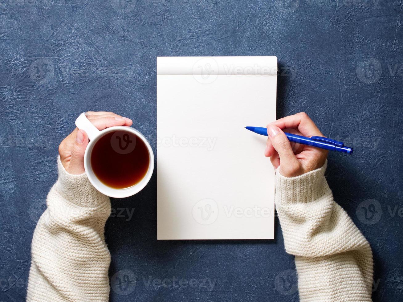 la donna scrive nel taccuino sul tavolo blu scuro, mano in camicia che tiene una matita, tazza di tè, disegno del quaderno di schizzi foto