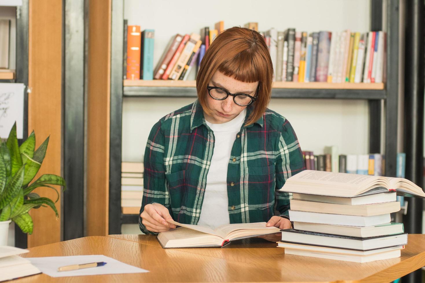 giovane donna rossa con gli occhiali legge il libro in biblioteca foto