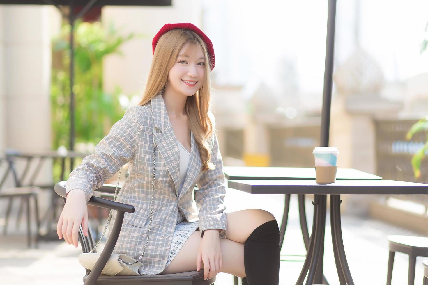 la giovane bella donna asiatica che indossa un abito e un berretto rosso con i capelli di bronzo si siede su una sedia nella caffetteria all'aperto mentre c'era una tazza di caffè sul tavolo in una mattina di sole. foto