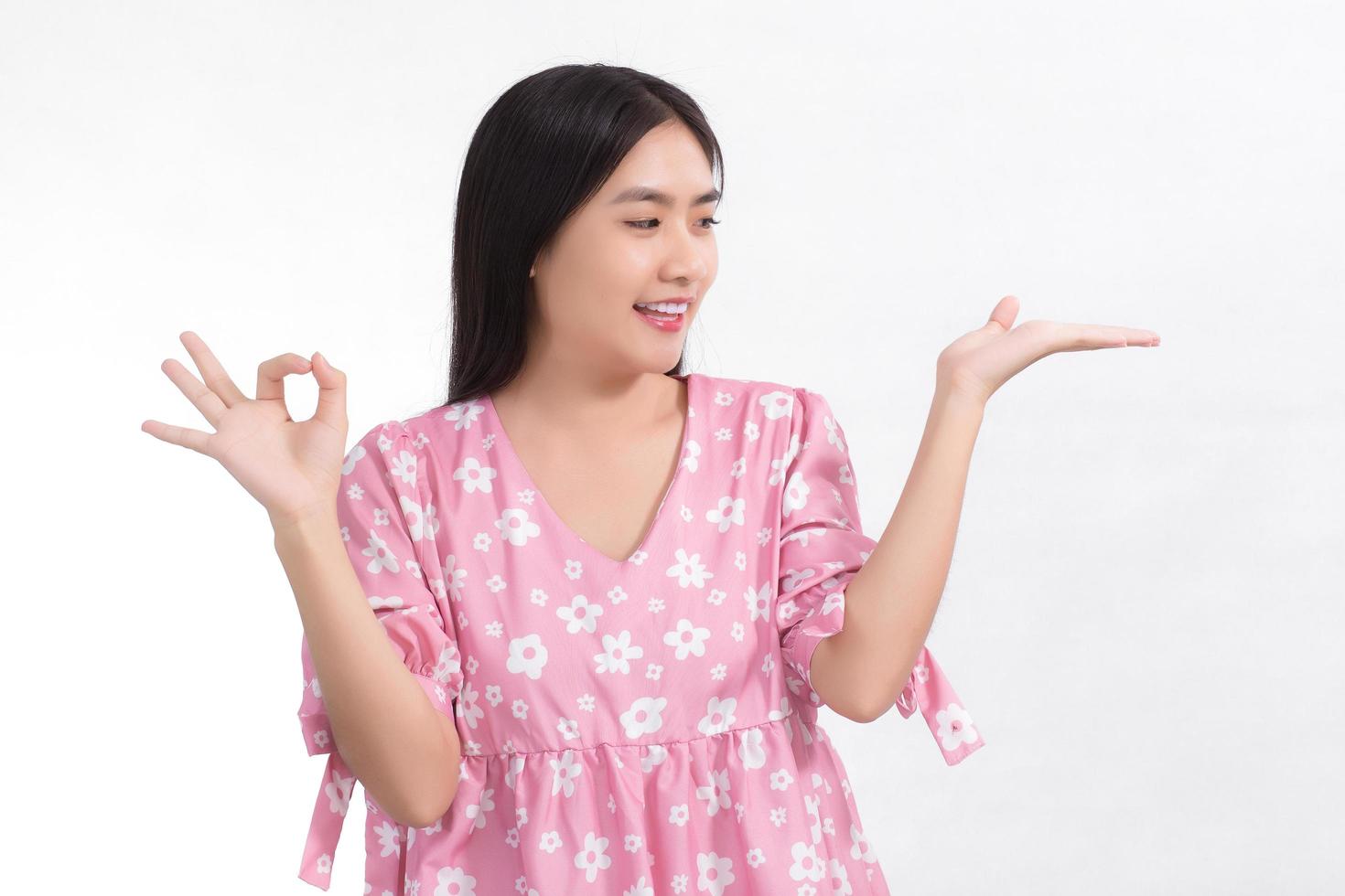 una donna carina asiatica dai capelli lunghi nera che indossa un abito rosa sta guardando la sua mano e mostra un segno okey su uno sfondo bianco. foto