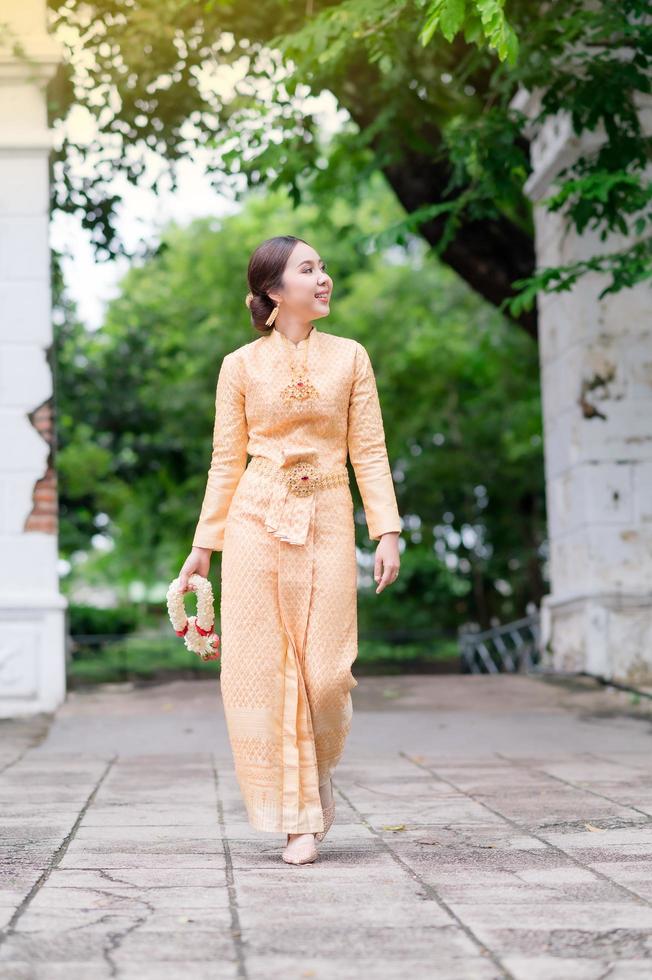 elegante donna tailandese in abito tailandese adornato con gioielli preziosi cammina tenendo ghirlande di fiori nel tempio tailandese foto
