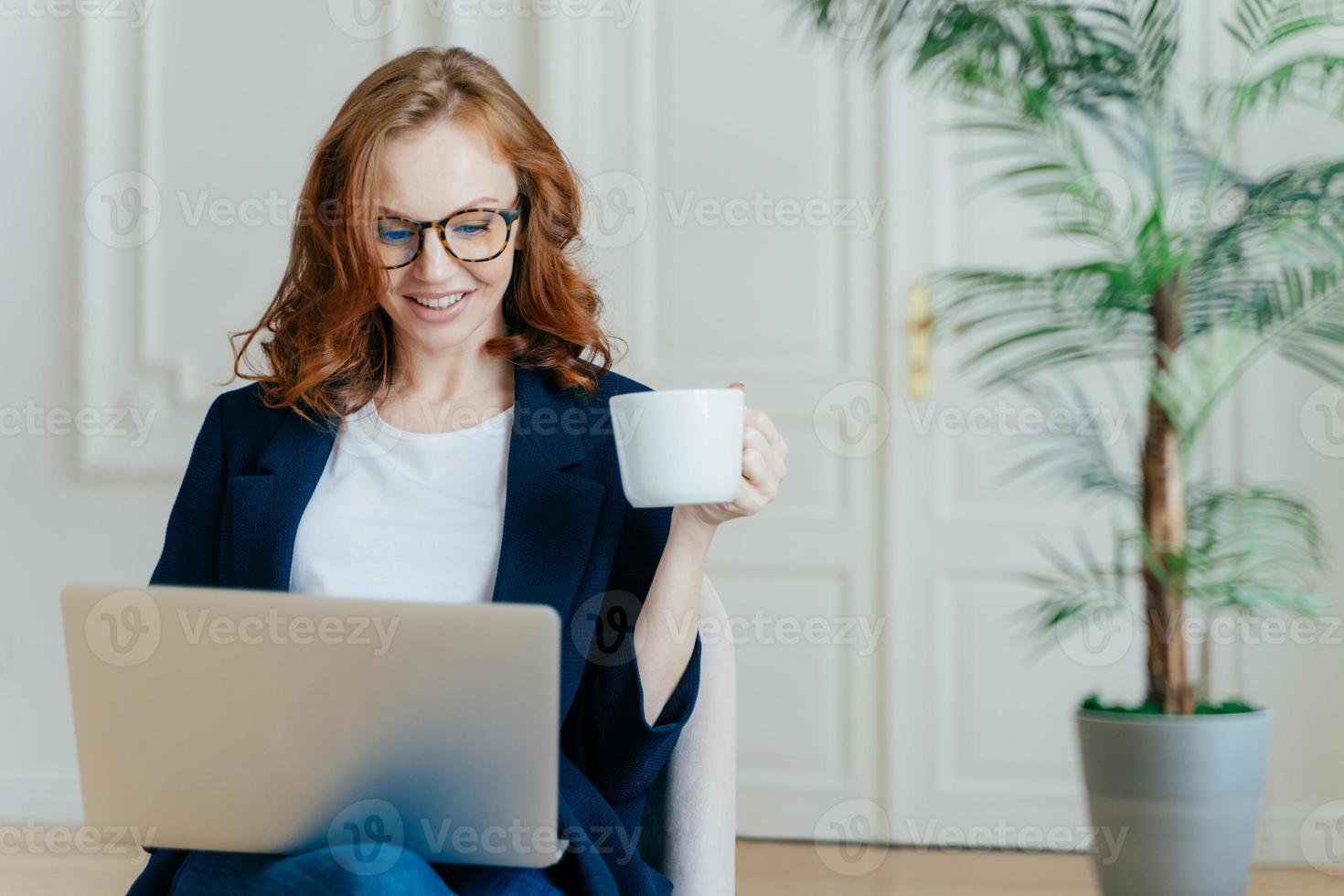 soddisfatta femmina europea con i capelli rossi, digita informazioni sul computer portatile, ha un'espressione facciale felice, beve caffè, indossa occhiali, sviluppa un nuovo progetto, posa da sola in una stanza accogliente. foto