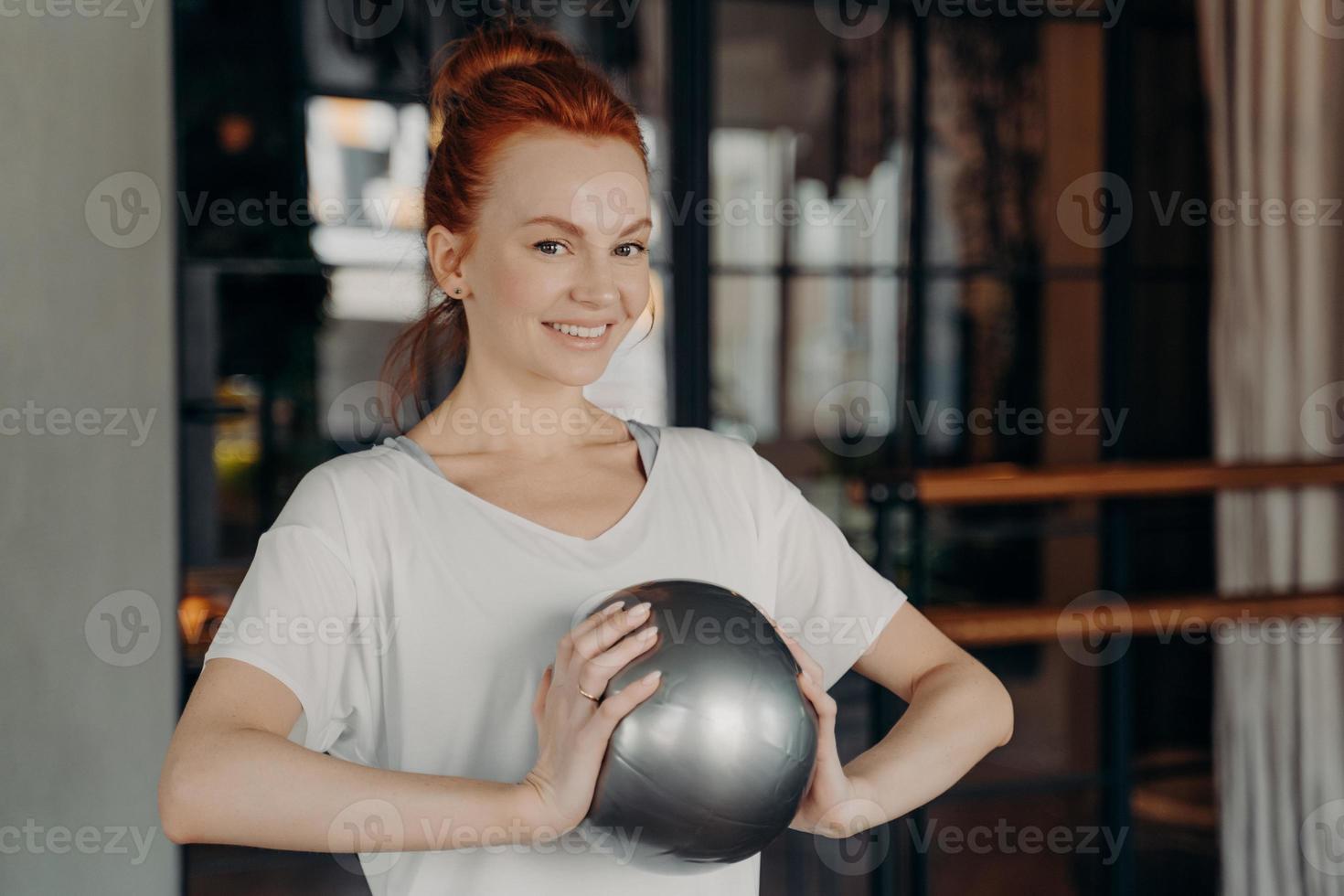 bella donna zenzero in abiti sportivi che sorride alla macchina fotografica mentre tiene in mano fitball argento di piccole dimensioni foto