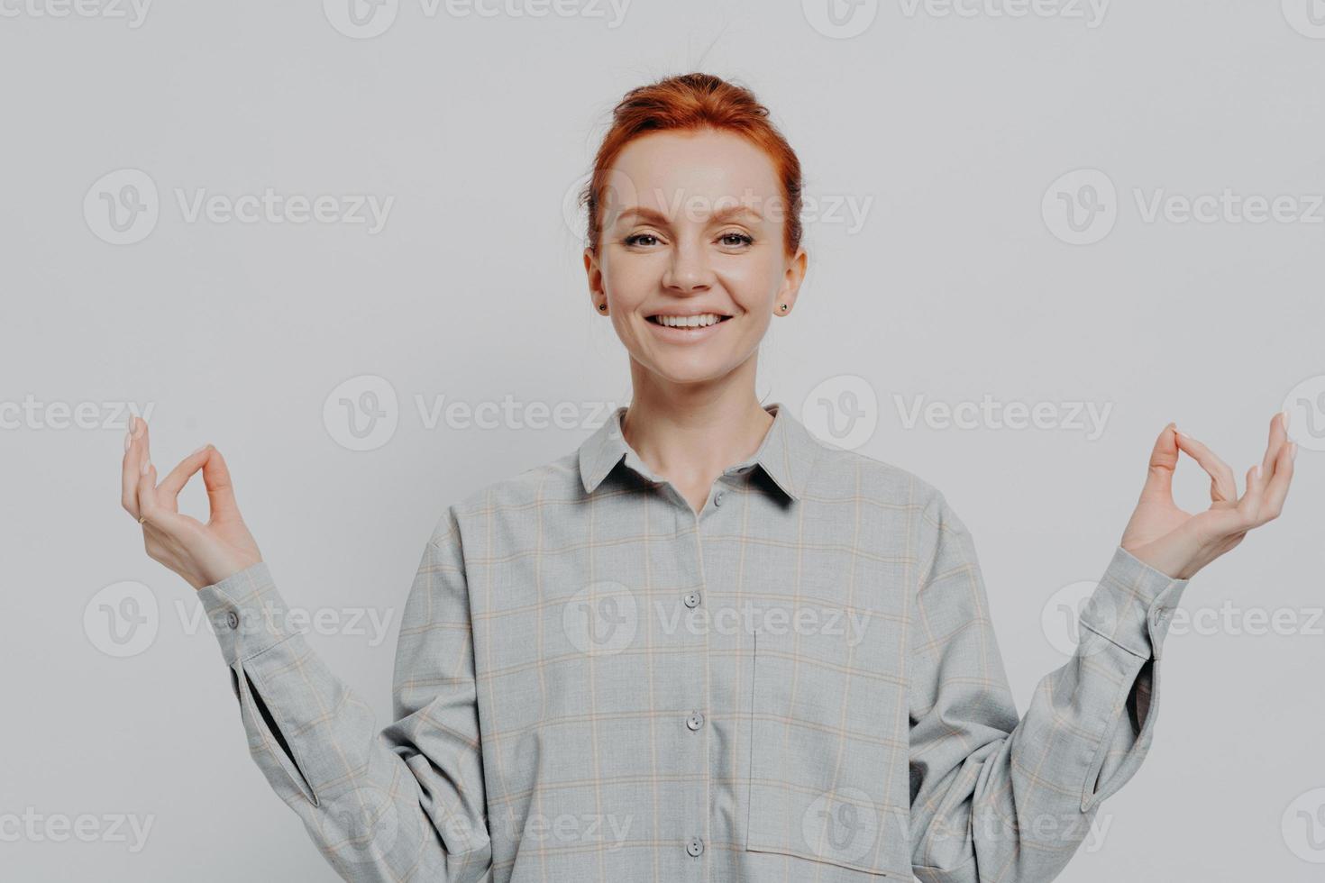giovane e pacifica donna rossa sorridente che si tiene per mano in mudra isolato su sfondo grigio per studio foto