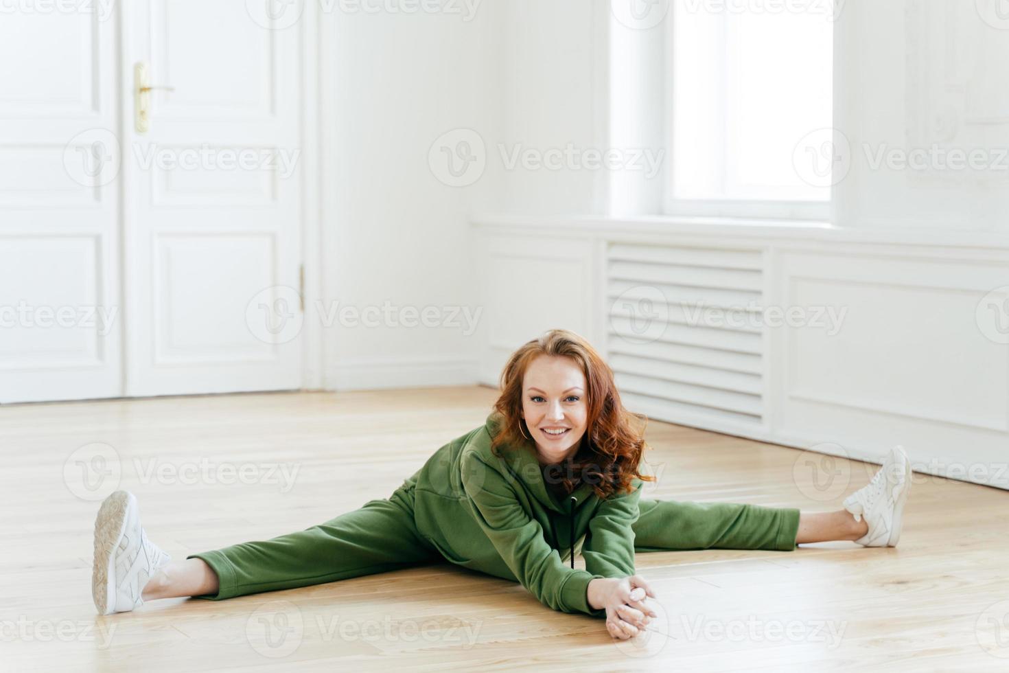 ginnasta in abbigliamento sportivo, fa la spaccatura delle gambe, si siede sul pavimento, posa nella sala yoga, fa esercizi di allenamento, allunga le gambe, dimostra le spaccate, gli piace il fitness. atleta donna attiva pone al coperto. foto