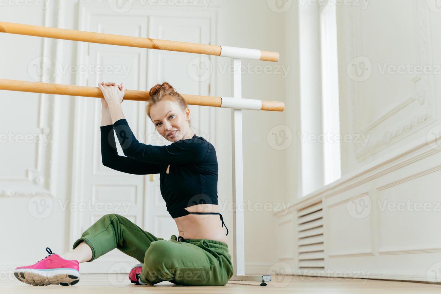 inquadratura di una bella donna rossa fa esercizi di pilates sul corrimano, si siede sul pavimento, si allunga, posa in uno studio di danza, è in buona forma fisica, vestita con abbigliamento sportivo. sport e assistenza sanitaria foto