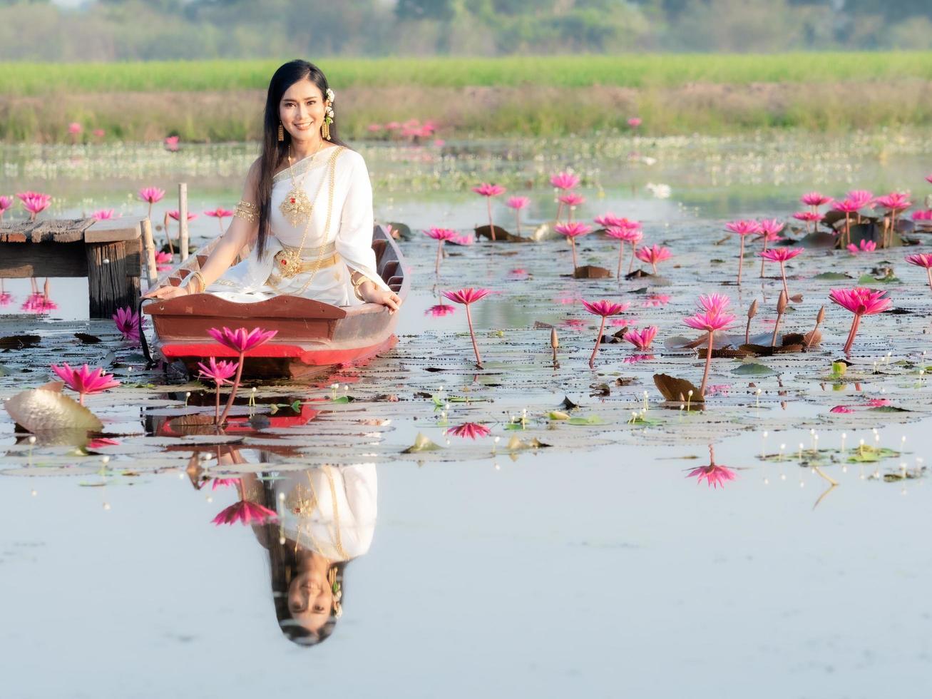 un'elegante donna tailandese che indossa abiti tradizionali tailandesi adornati con ornamenti d'oro, seduta su una barca di legno che galleggia in un campo di loto foto