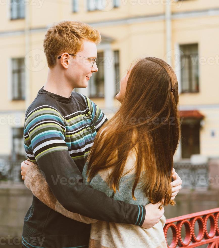 ragazza con una lunga e spessa udienza scura che abbraccia un ragazzo dai capelli rossi con la maglietta blu su un ponte, giovane coppia. concetto di amore adolescenziale e primo bacio, sentimenti sinceri di uomo e donna, città, lungomare foto