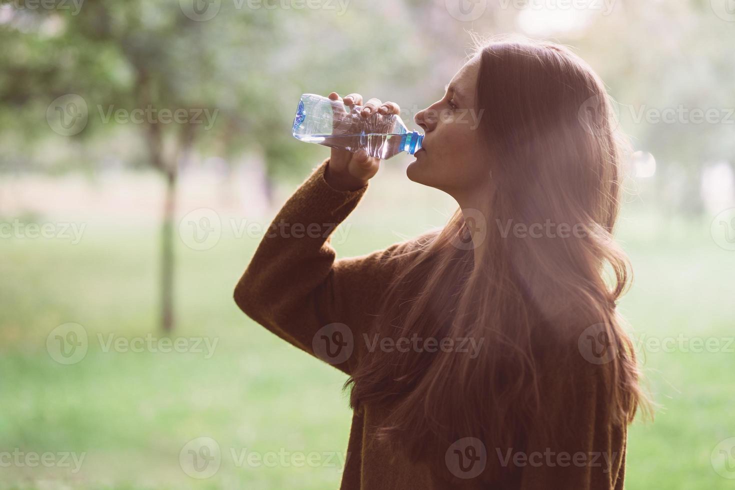giovane bella ragazza che beve acqua da una bottiglia di plastica sulla strada nel parco in autunno o in inverno. una donna con bei capelli lunghi e folti scuri disseta la sua sete di acqua durante una passeggiata foto
