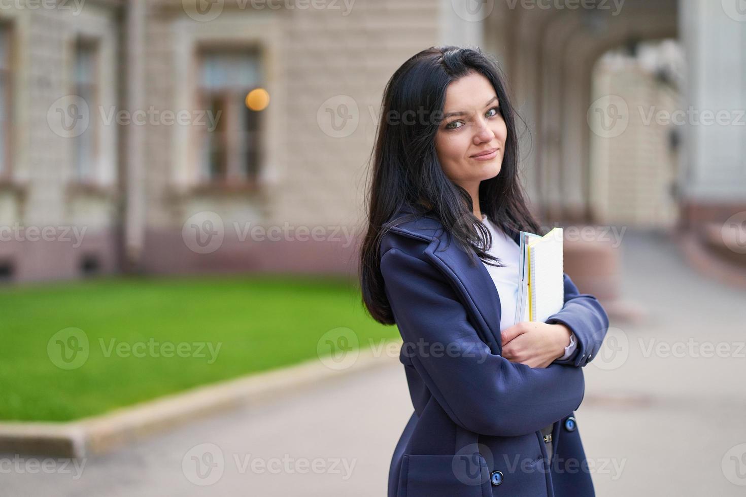 sorridente ragazza bruna studente in possesso di quaderni e libri di testo, si trova all'università sulla strada di st. pietroburgo. una donna affascinante con lunghi capelli scuri sta studiando al corso, ricevendo istruzione foto