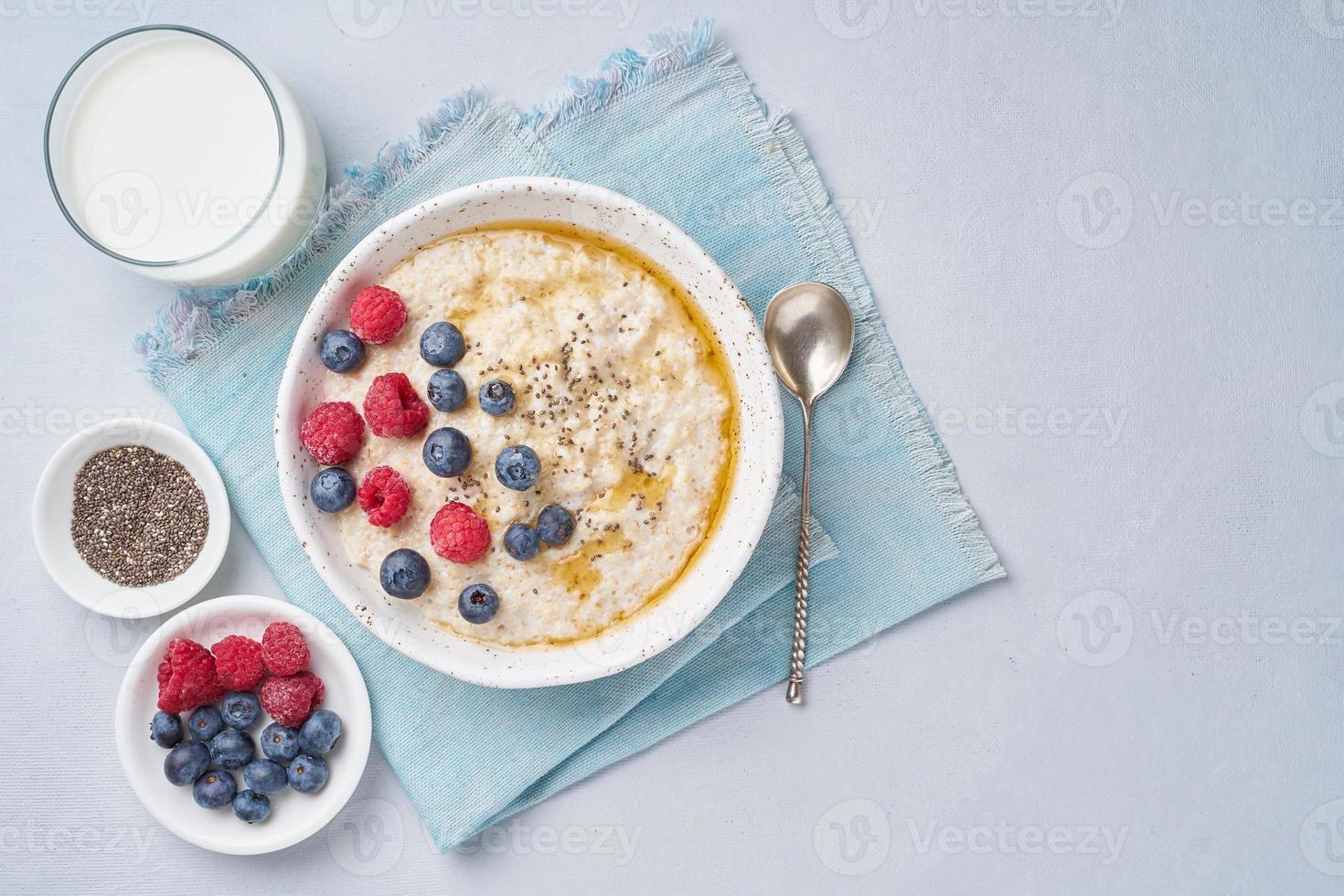 farina d'avena con frutti di bosco, chia, sciroppo d'acero e bicchiere di latte su sfondo blu chiaro. vista dall'alto, copia spazio. colazione dietetica sana foto