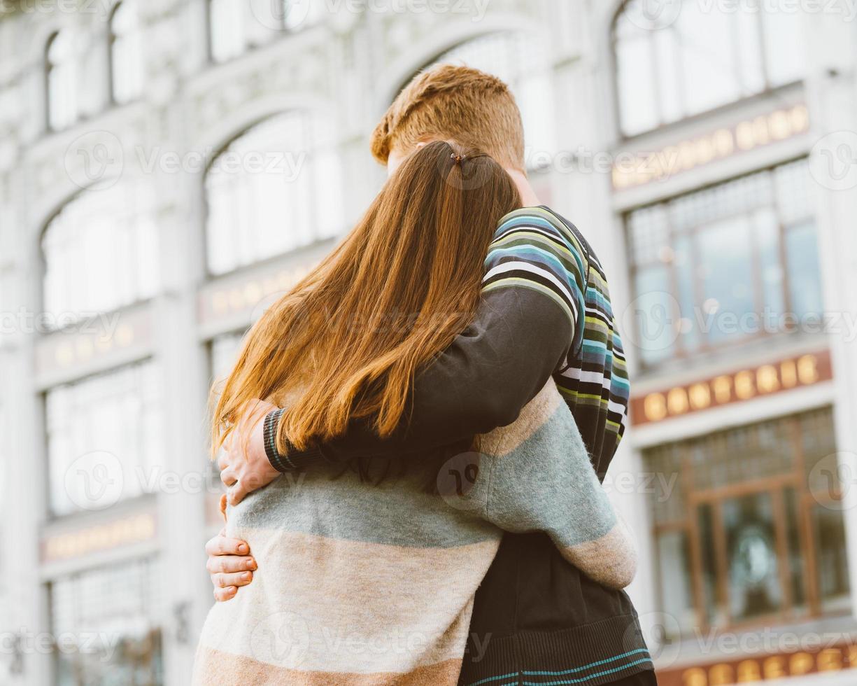 ragazza con lunghi capelli scuri e folti che si tengono per mano ragazzo dai capelli rossi in maglietta blu sul ponte, amore per adolescenti la sera. ragazzo guarda teneramente ragazza, giovane coppia. concetto di amore adolescenziale e primo bacio foto