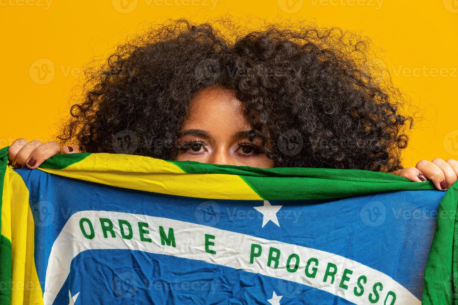 misteriosa fan della donna di colore che tiene in faccia una bandiera brasiliana. colori brasiliani sullo sfondo, verde, blu e giallo. elezioni, calcio o politica. foto