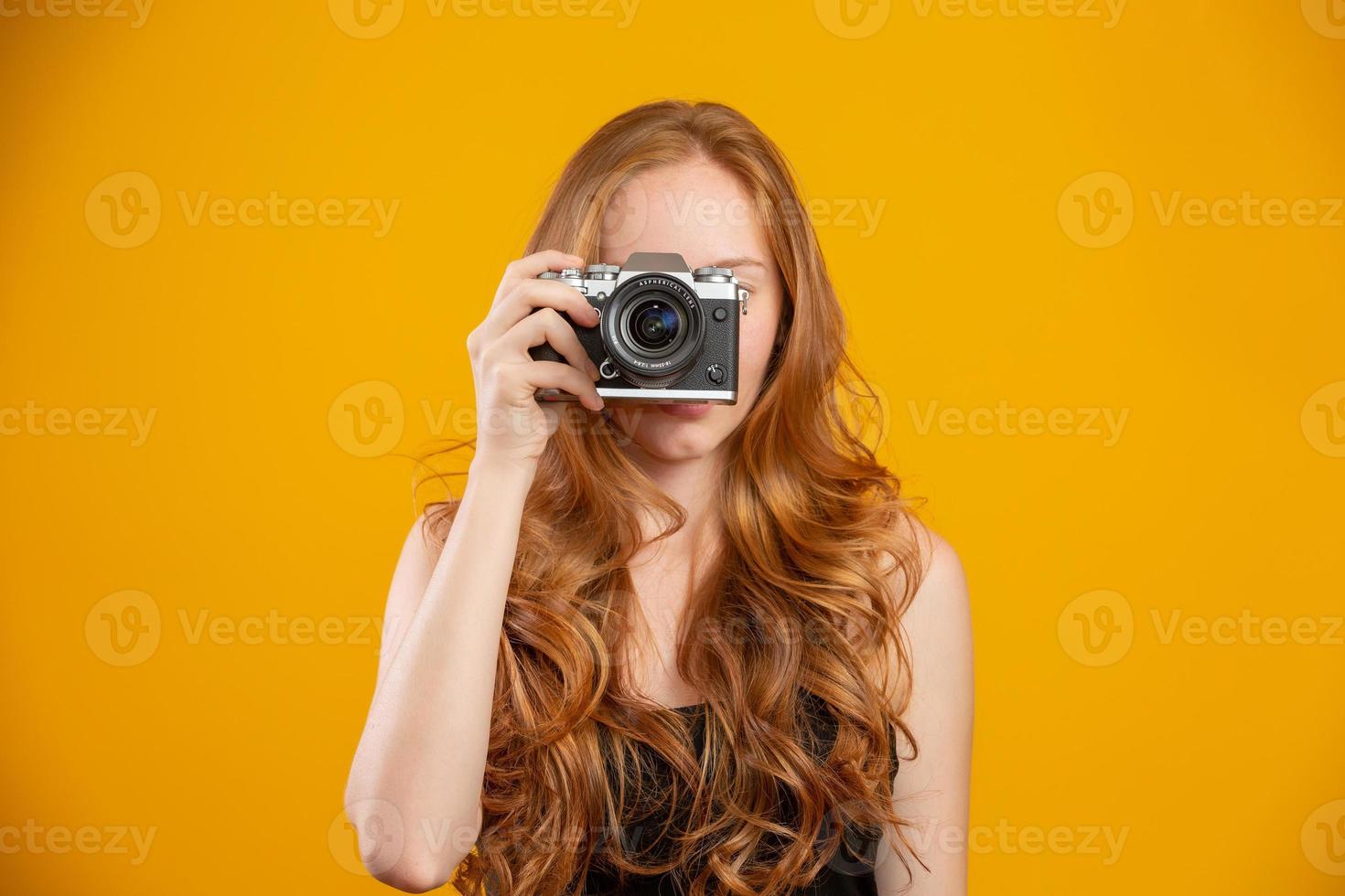 foto di adorabile donna rossa che indossa abiti neri che tiene una fotocamera vintage retrò e scatta foto isolate su sfondo giallo. simulare lo spazio di copia. tenere una macchina fotografica vintage retrò.