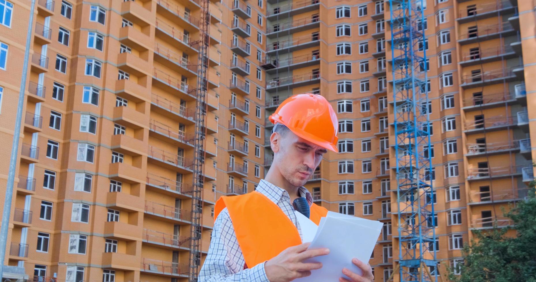 ritratto di specialista edile in casco arancione e giubbotto di sicurezza contro il grande edificio foto