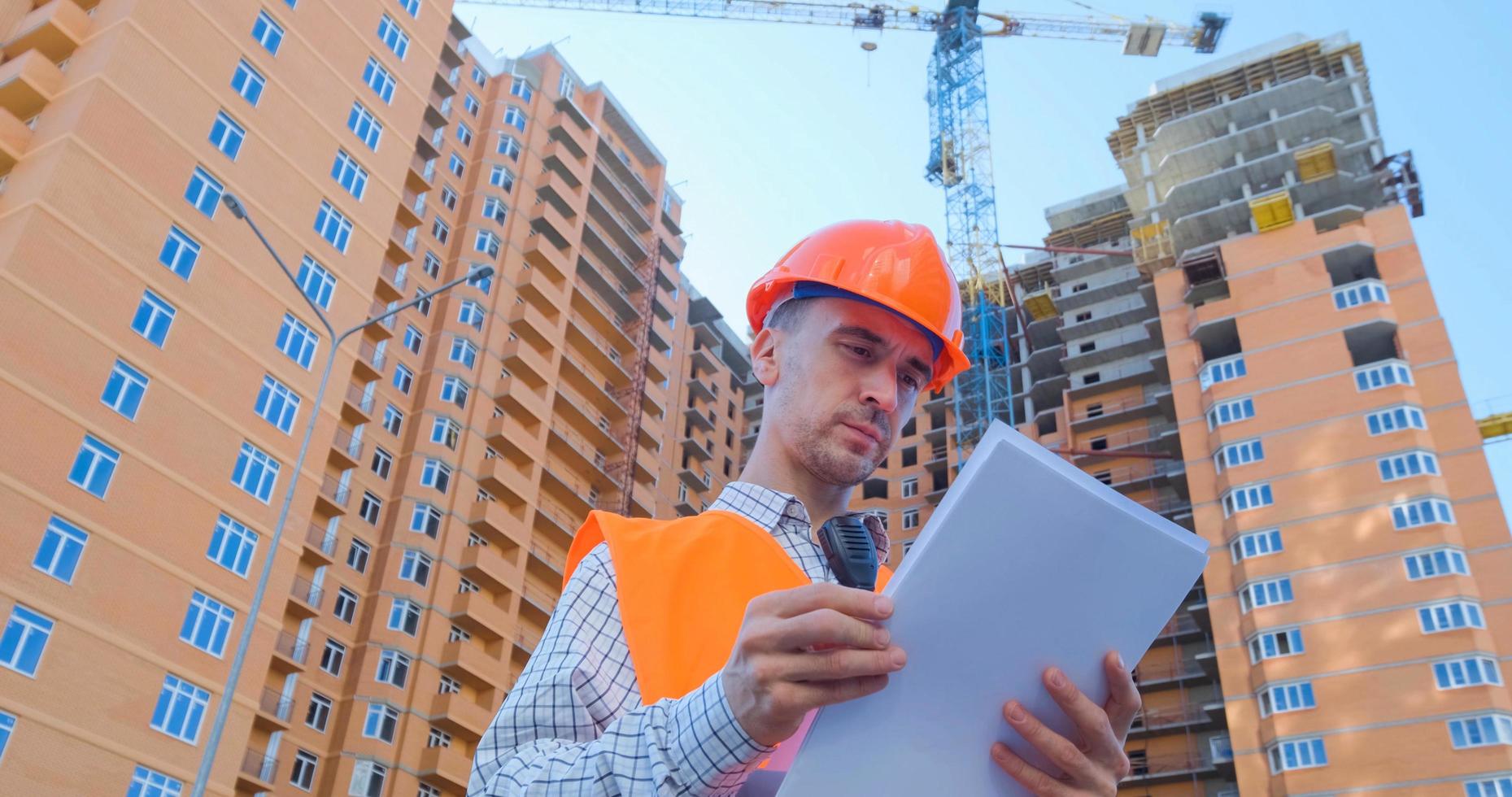 ritratto di specialista edile in casco arancione e giubbotto di sicurezza contro il grande edificio foto