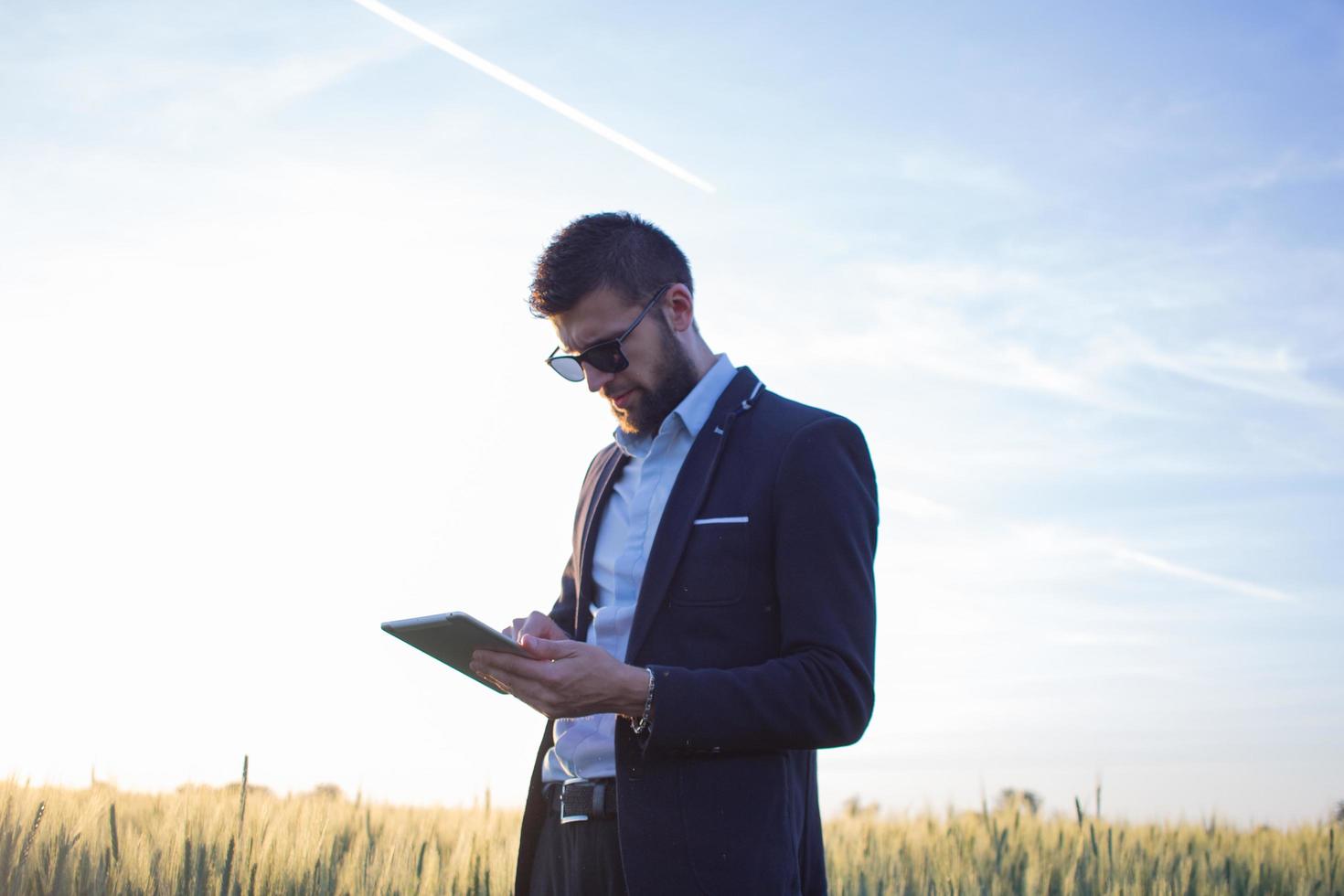 uomo d'affari in campi di grano estivi utilizzando tablet durante il tramonto, uomo in tuta con computer compatto foto