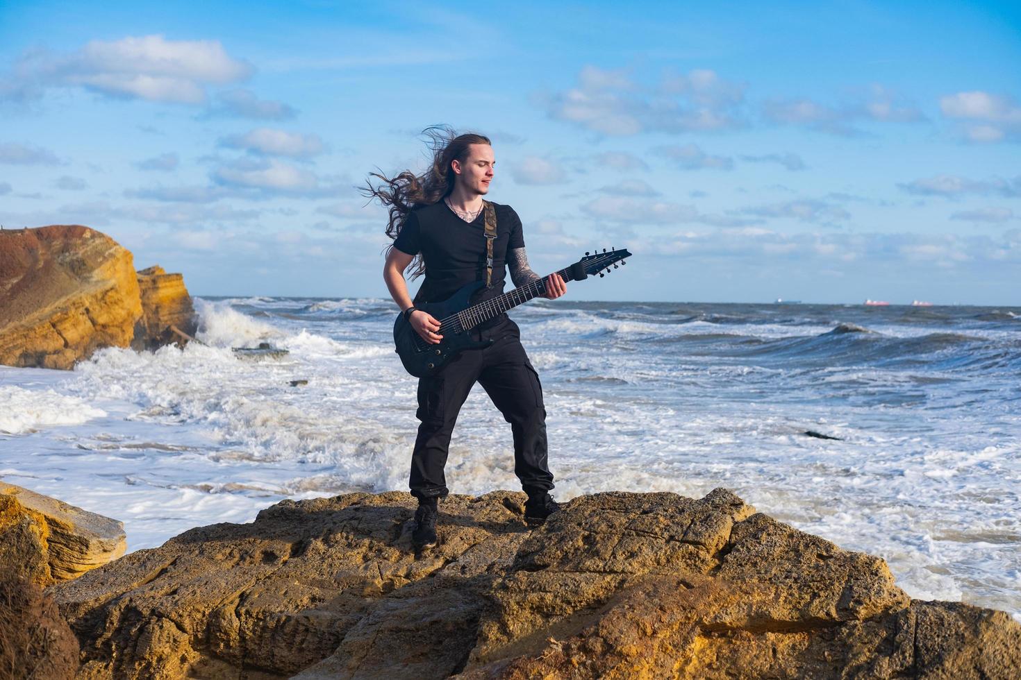 giovane maschio in nero e capelli lunghi suona la chitarra sulla spiaggia foto