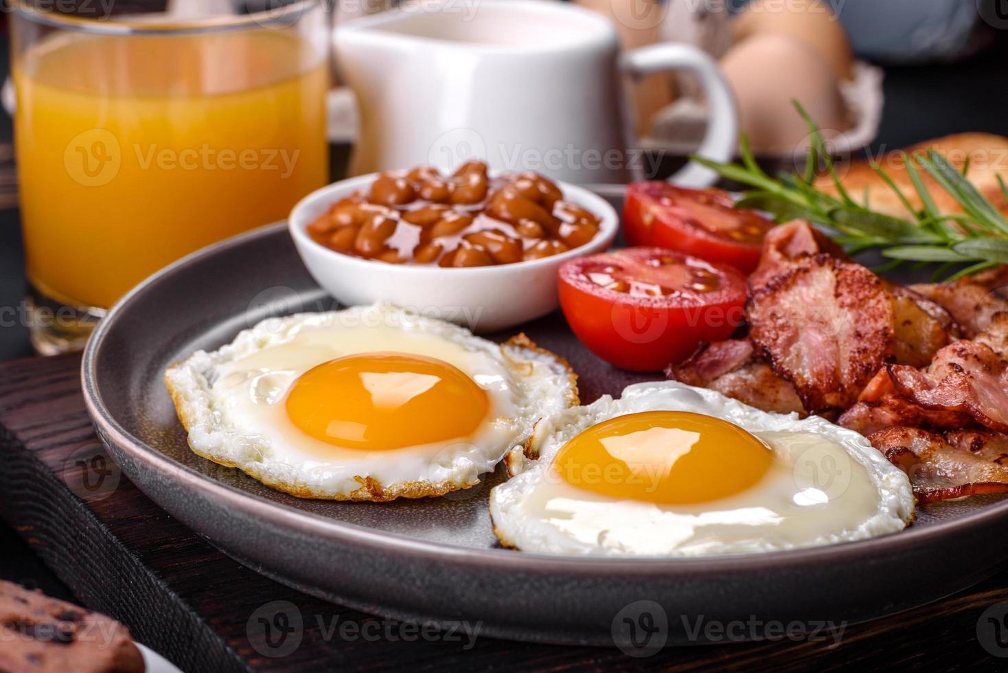 una deliziosa colazione sana e nutriente con uova fritte, pancetta, fagioli, un bicchiere di succo, biscotti d'avena, latte e marmellata foto