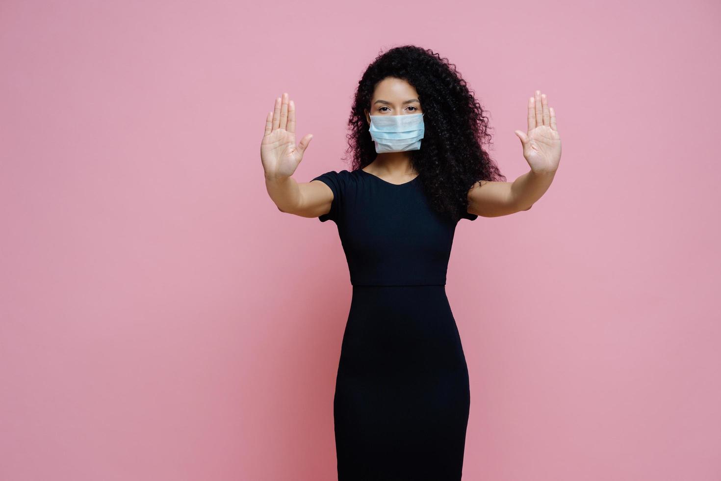 l'immagine di una seria donna afroamericana tira i palmi verso la telecamera, fa un gesto di arresto, un segno di rifiuto, previene il coronavirus o una malattia contagiosa, indossa una maschera usa e getta, isolata su sfondo rosa foto