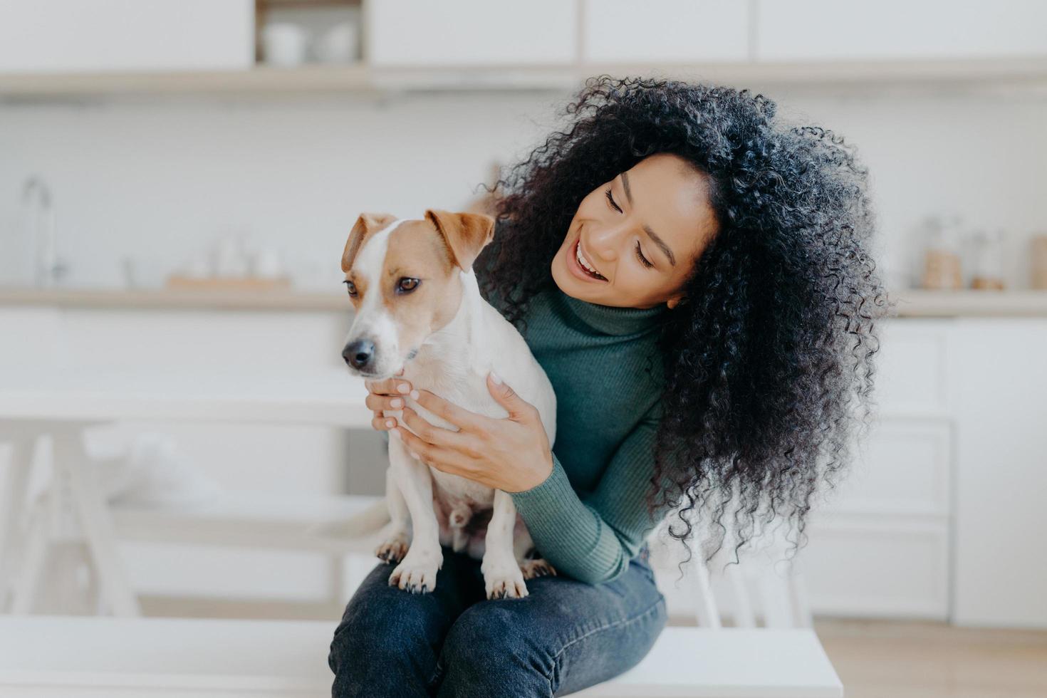 felice proprietaria del cane jack russell terrier, si sente responsabile della cura dell'animale domestico, ha folti capelli ricci scuri, si siede su uno sfondo sfocato della cucina. persone e rapporto con gli animali foto