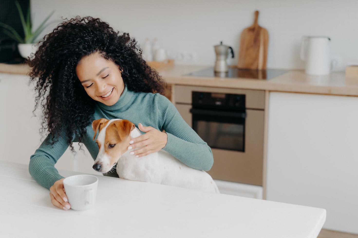 felice donna afro africana con l'acconciatura riccia tratta il cane in cucina, posa al tavolo bianco con una tazza di bevanda, goditi l'atmosfera domestica, fai colazione insieme. persone, animali, concetto di casa. foto