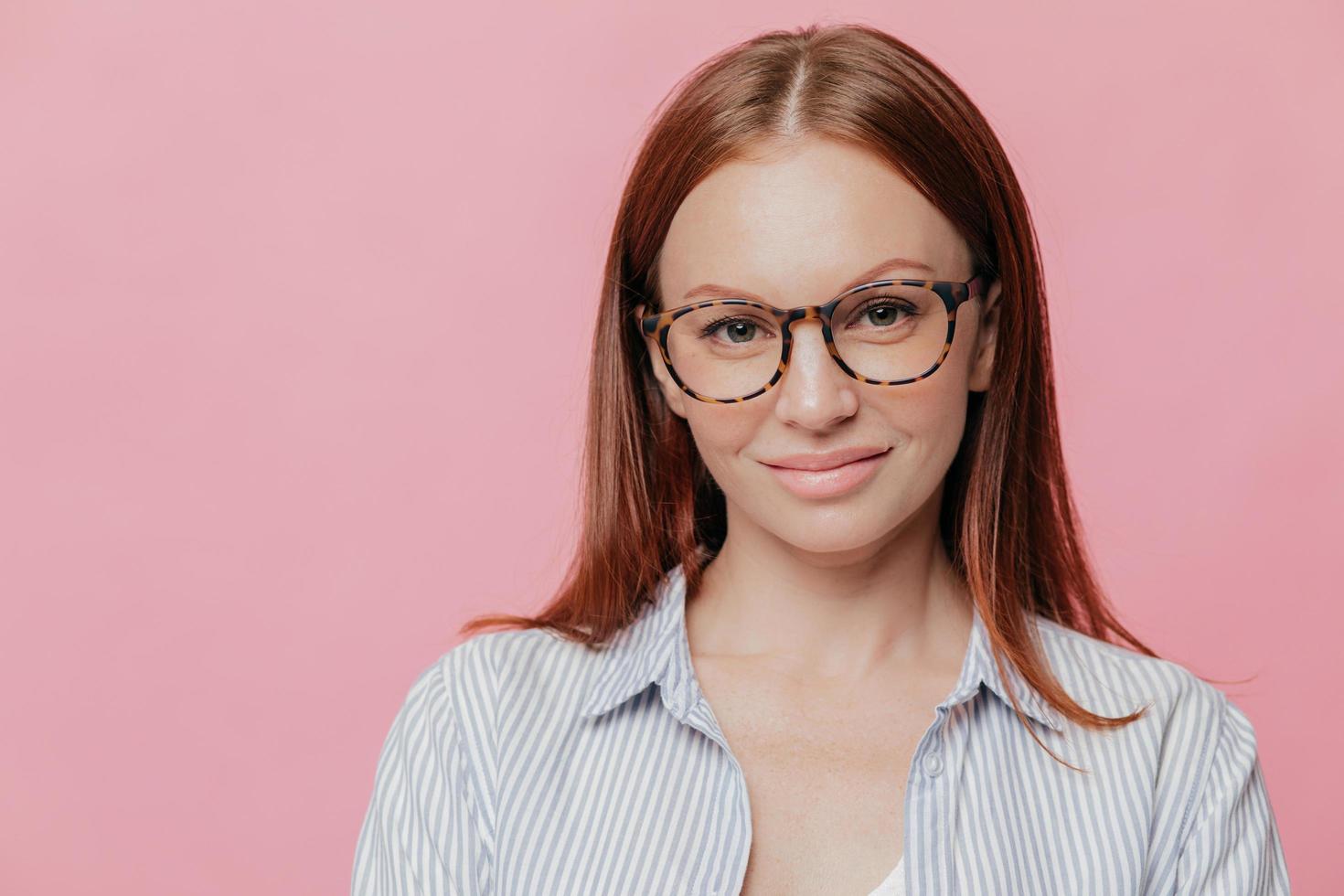 la giornalista soddisfatta indossa una camicia elegante, occhiali da vista, ha le labbra carnose, è felice di incontrare persone, posa contro il muro rosa dello studio con uno spazio vuoto per i tuoi contenuti promozionali. tiro al coperto foto