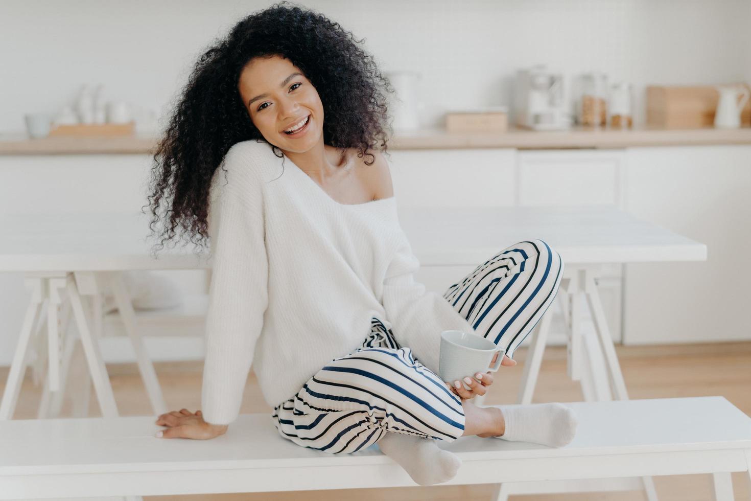 giovane ragazza soddisfatta con acconciatura afro riccia, siede a gambe incrociate sulla panchina, gode del caffè mattutino e dell'atmosfera domestica, posa sullo sfondo della cucina, trascorre del tempo da solo, fa colazione foto