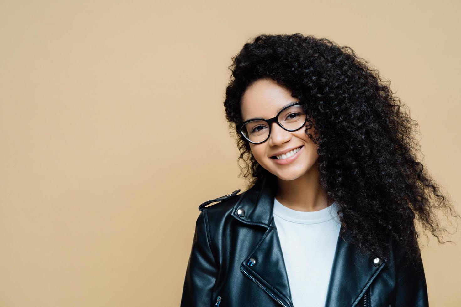 inquadratura orizzontale di donna sana e felice con capelli ricci naturali, sorriso piacevole, gode del tempo libero e fa shopping nel fine settimana, indossa occhiali trasparenti, giacca di pelle, isolato su sfondo beige foto