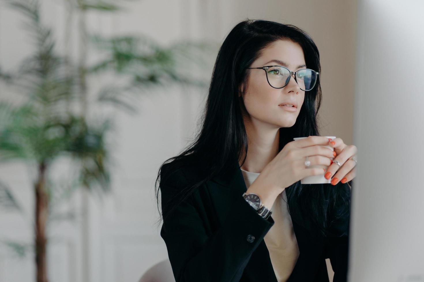 foto di una copywriter bruna concentrata beve caffè, lavora su compiti creativi, indossa occhiali e costume nero formale, guarda webinar per migliorare le abilità, posa nello spazio di coworking