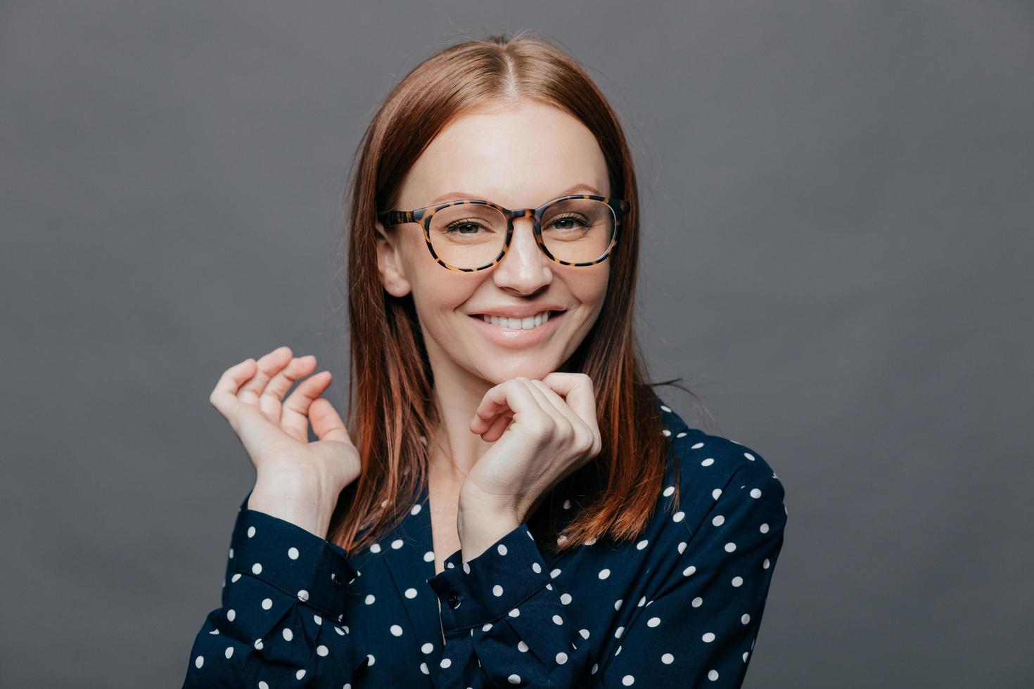 positiva attraente donna caucasica tiene la mano vicino al mento, guarda positivamente la fotocamera, tiene la mano sotto il mento, sorride felicemente, indossa una camicia nera con pois bianchi, posa su sfondo grigio. foto