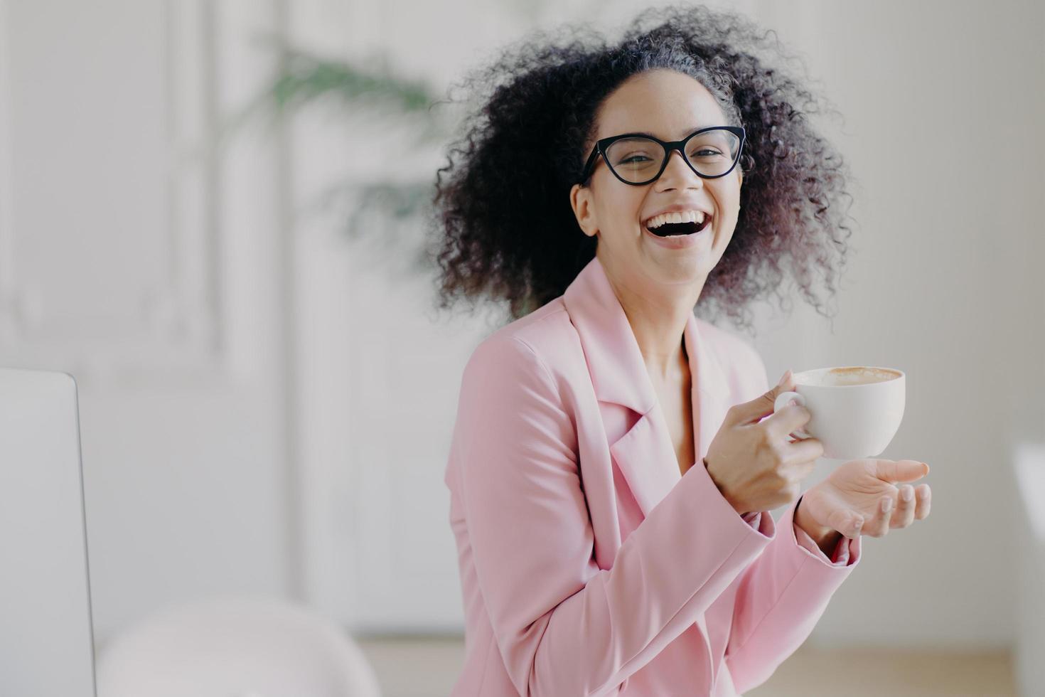 la donna dai capelli ricci felicissima ride allegramente mentre beve caffè caldo o latte macchiato, indossa occhiali trasparenti, si diverte durante la pausa in ufficio, mostra i denti bianchi, è un'imprenditrice professionista foto