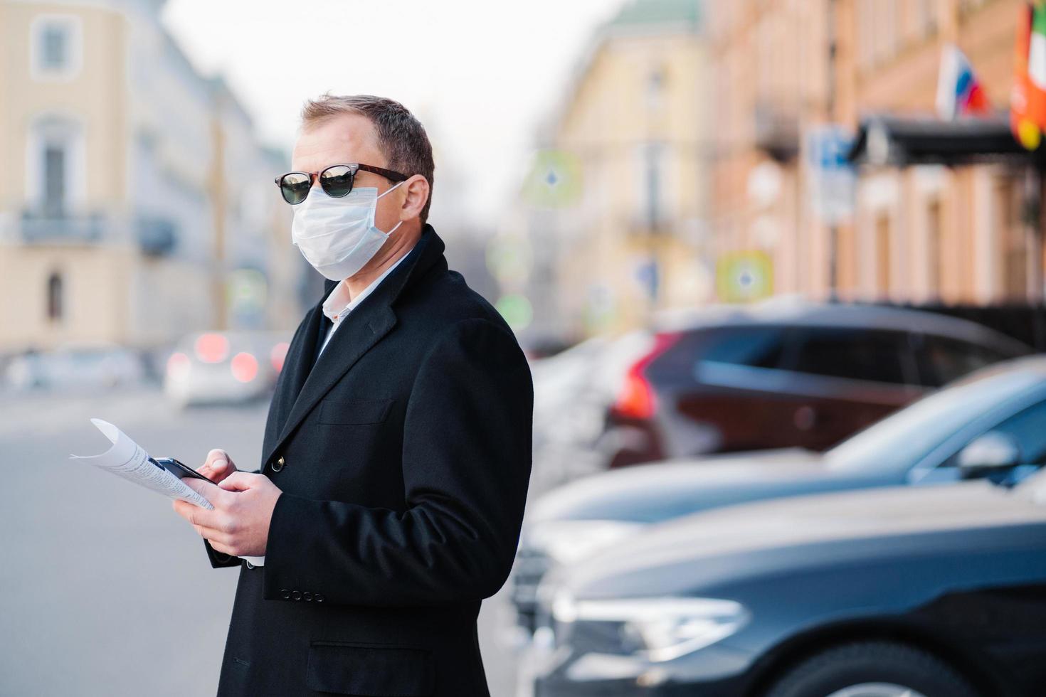 focolaio di coronavirus pandemico. un uomo d'affari serio posa all'aperto vicino ai trasporti in strada, tiene cellulari e giornali moderni, vestito con un cappotto nero, indossa una maschera protettiva dal coronavirus. foto