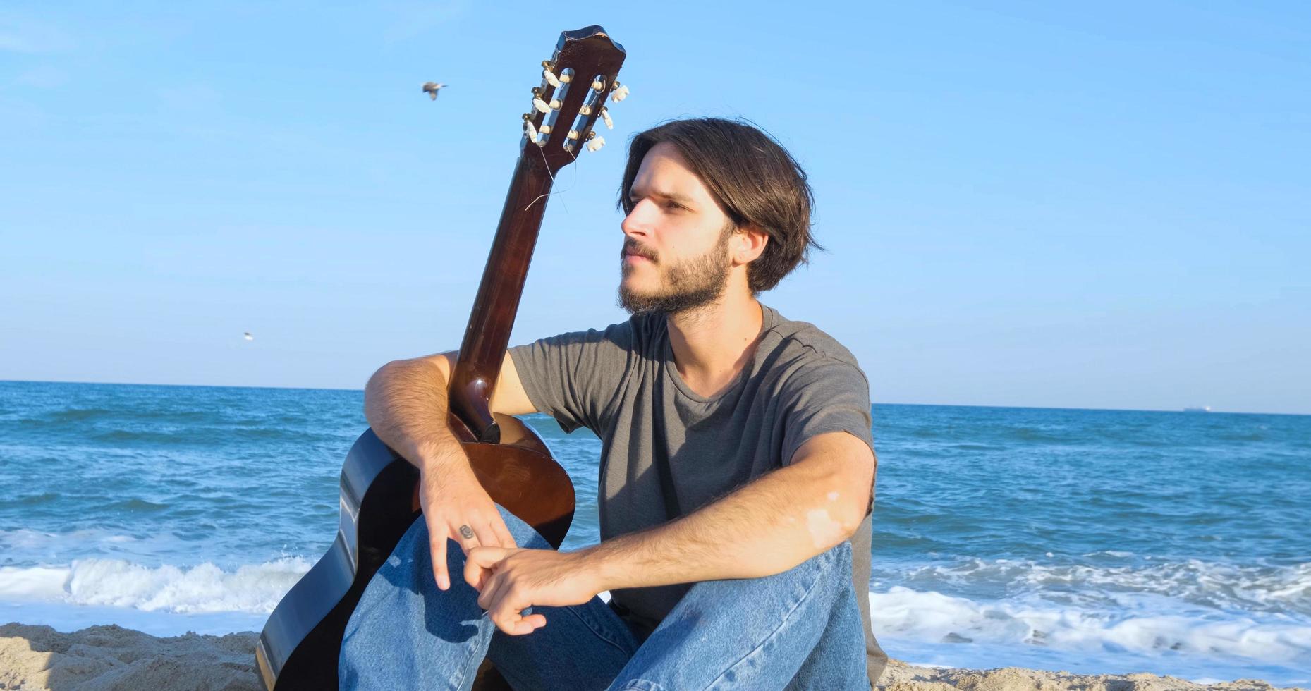 giovane maschio bello suonare in chitarra acustica sulla spiaggia in una giornata di sole, mare o oceano sullo sfondo foto