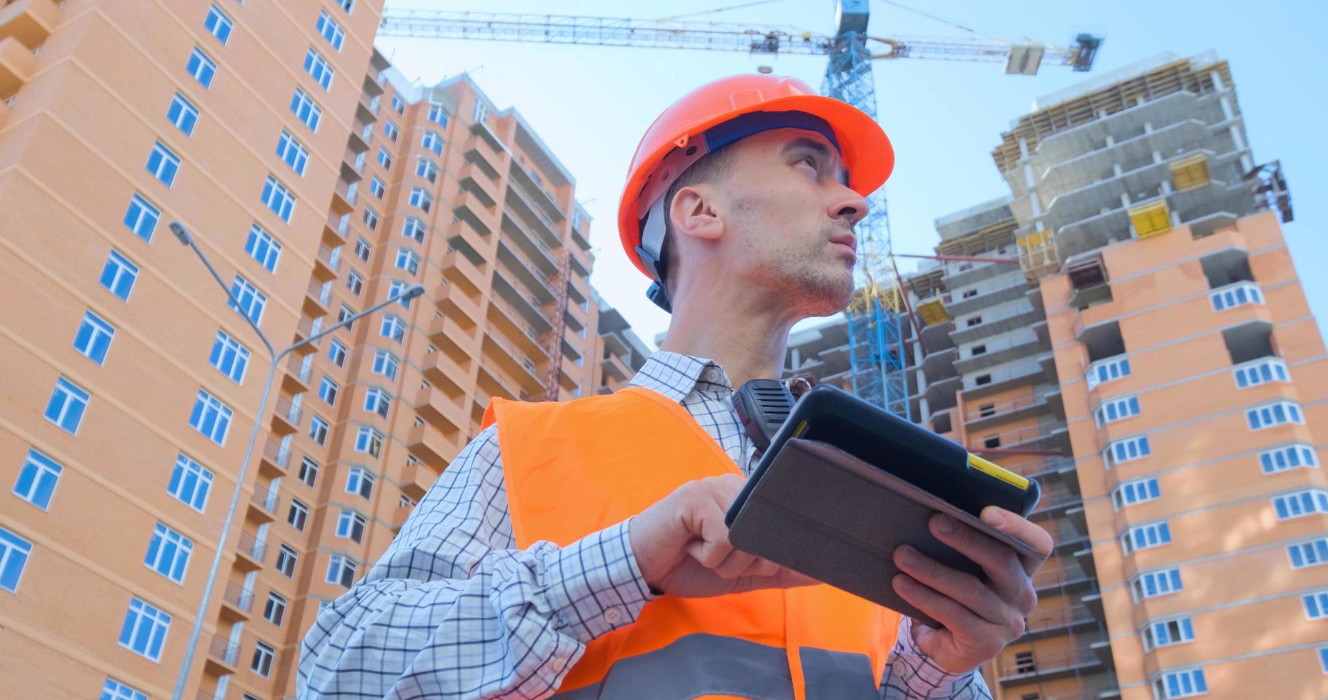 ritratto di specialista edile in casco arancione e giubbotto di sicurezza contro il grande edificio foto