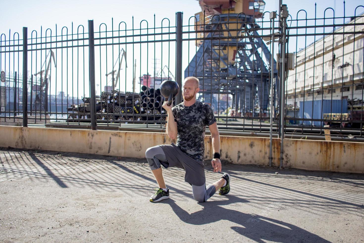 giovane atleta maschio barbuto che si allena nella zona industriale in una giornata di sole, esercizi di kettlebell all'aperto, fondo urbano foto