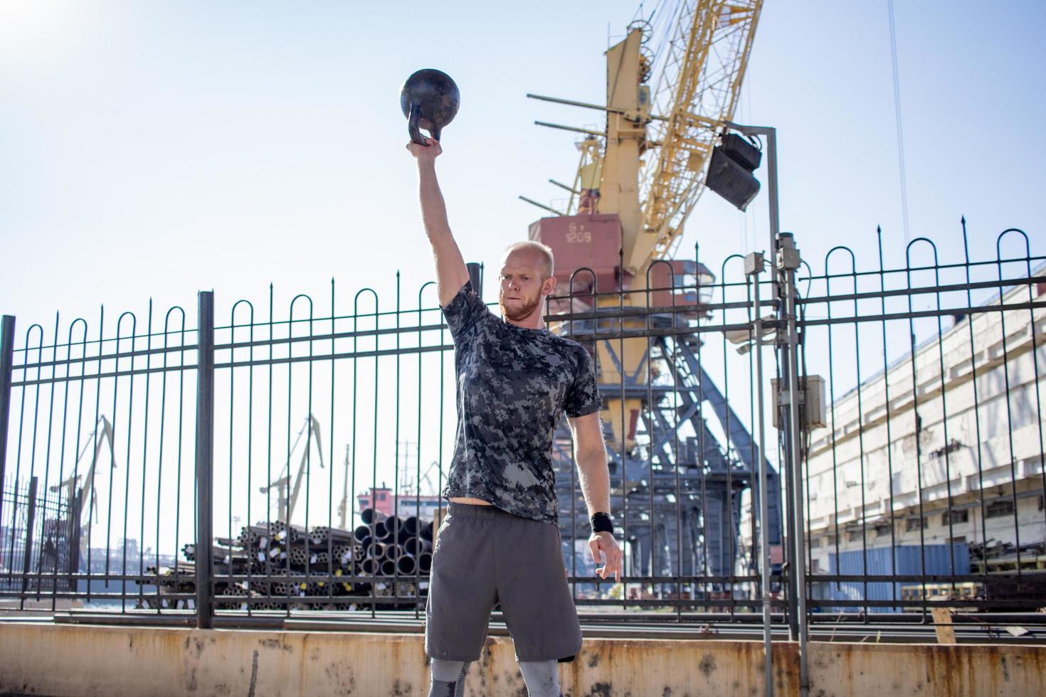 giovane atleta maschio barbuto che si allena nella zona industriale in una giornata di sole, esercizi di kettlebell all'aperto, fondo urbano foto