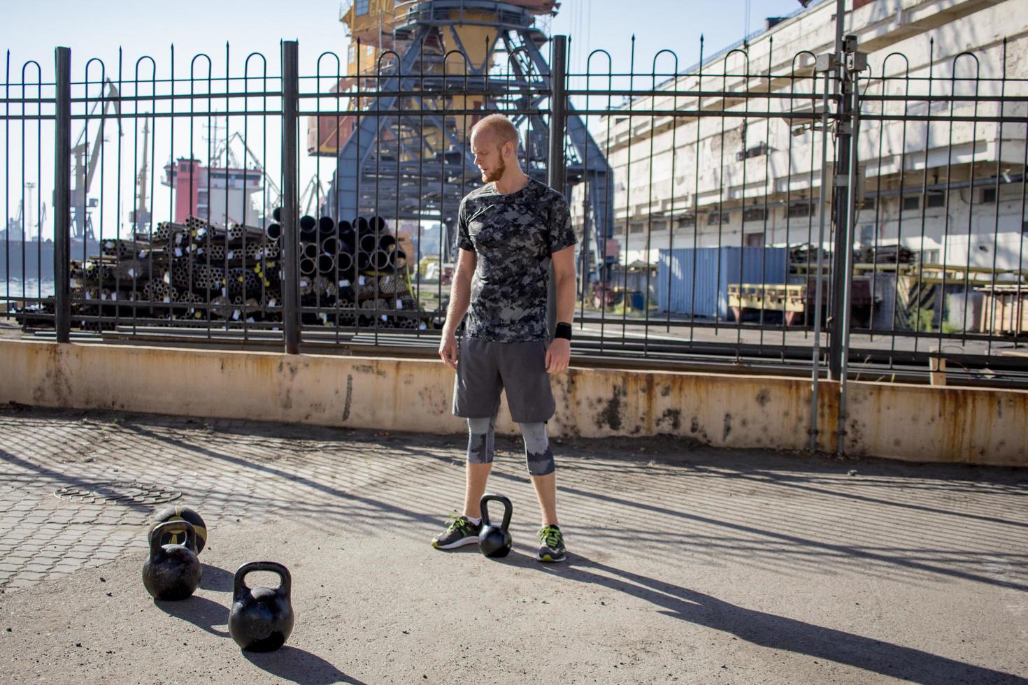 giovane atleta maschio barbuto che si allena nella zona industriale in una giornata di sole, esercizi di kettlebell all'aperto, fondo urbano foto