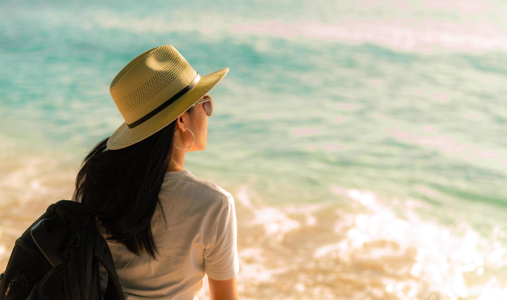 vista posteriore di felice giovane donna asiatica in stile casual con cappello di paglia e zaino. rilassati e goditi le vacanze sulla spiaggia del paradiso tropicale. vibrazioni estive. rilassarsi e godersi la spiaggia tropicale. foto