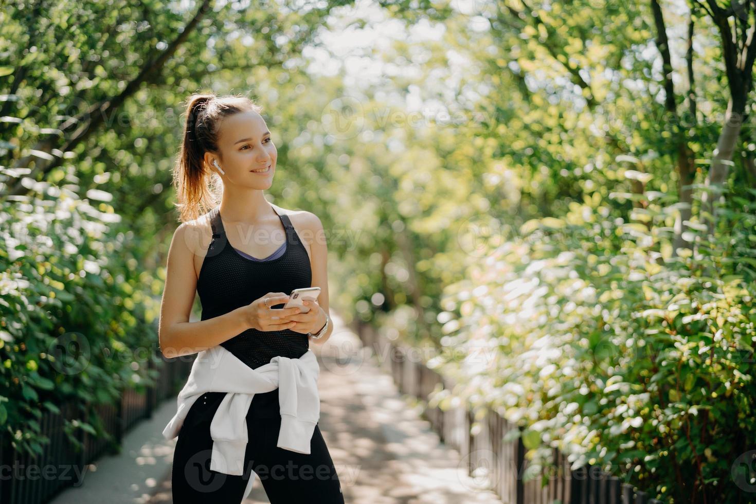 l'inquadratura orizzontale di una donna sportiva attiva utilizza lo smartphone per controllare i risultati dopo aver fatto jogging vestito con abbigliamento attivo gode di una calda giornata di sole ascolta musica tramite gli auricolari. concetto di stile di vita sano. foto