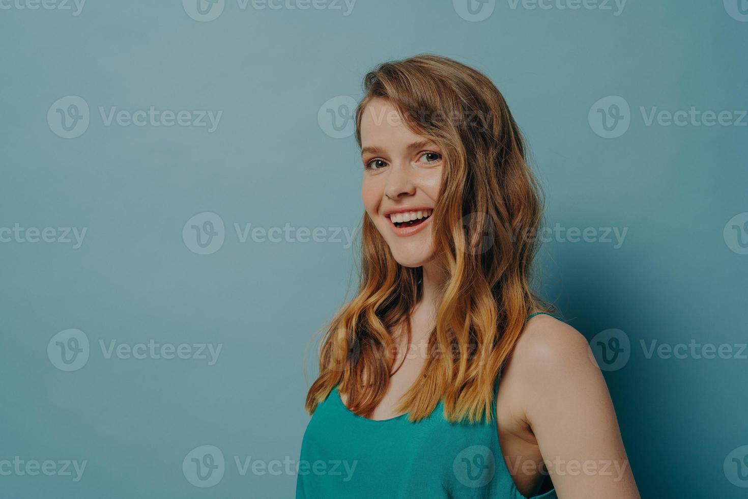 la ragazza millenaria positiva con i capelli ondulati sta di lato e guarda la fotocamera con un sorriso radioso e gioioso foto