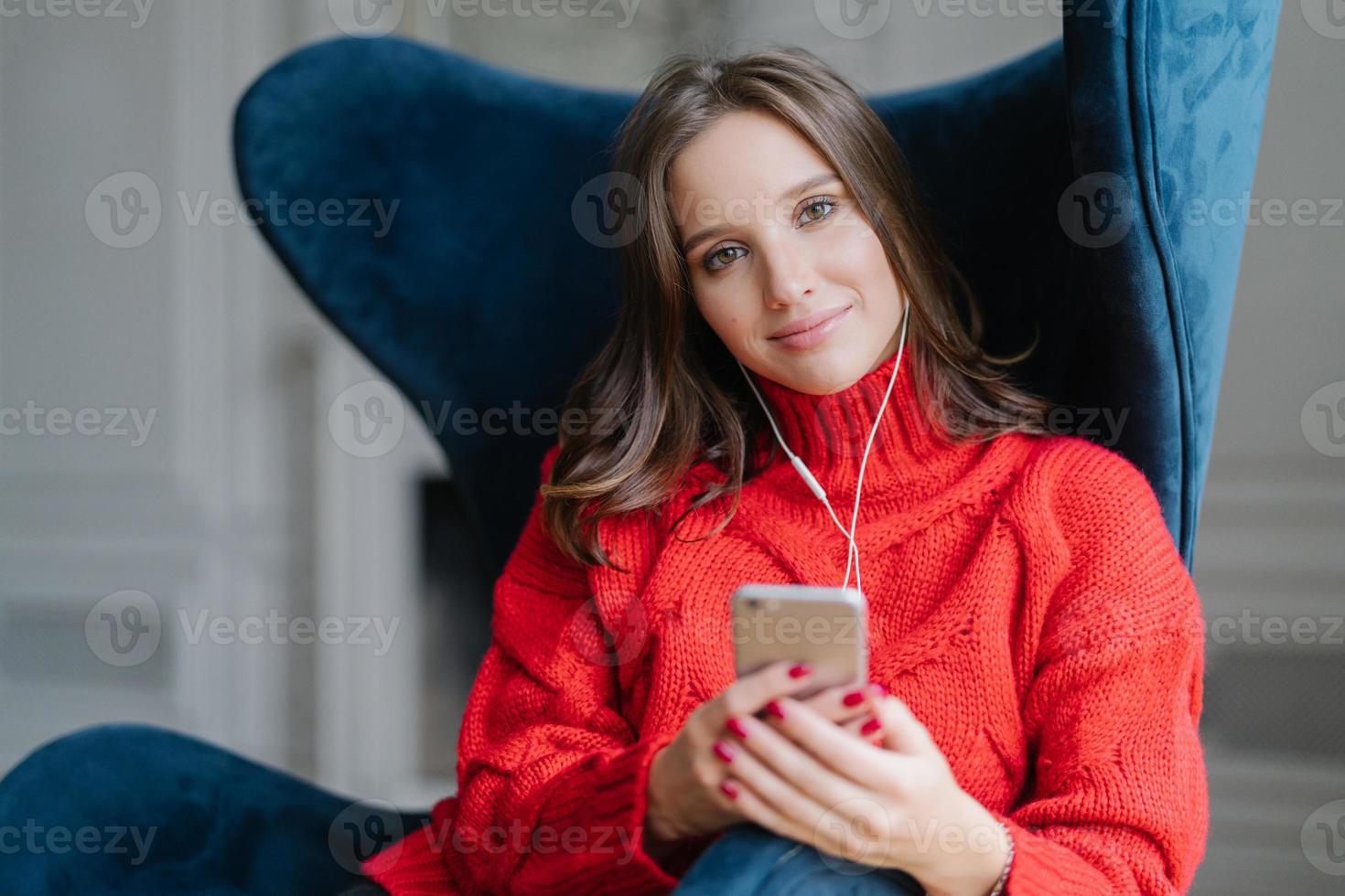 concetto di persone e tecnologia. La meloman femminile soddisfatta ama la musica preferita, ascolta registrazioni audio, vestita con un maglione rosso, naviga nella playlist, si siede in poltrona in soggiorno, ha tempo libero foto
