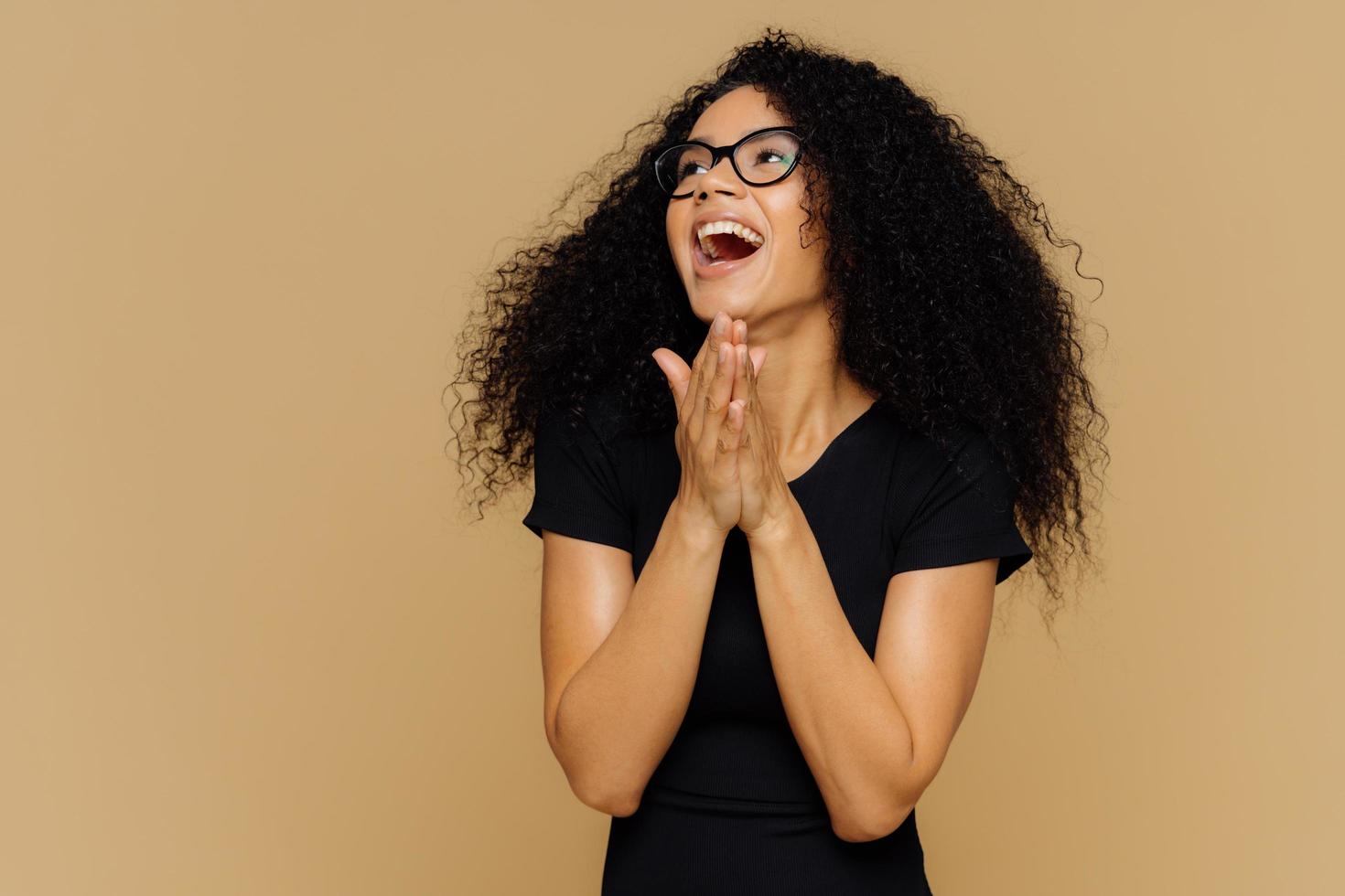 bella donna felicissima stringe le mani, ride gioiosamente, distoglie lo sguardo, si concentra da parte, esprime felicità, ha i capelli ricci folti, indossa abiti casual, occhiali, isolata su sfondo marrone foto