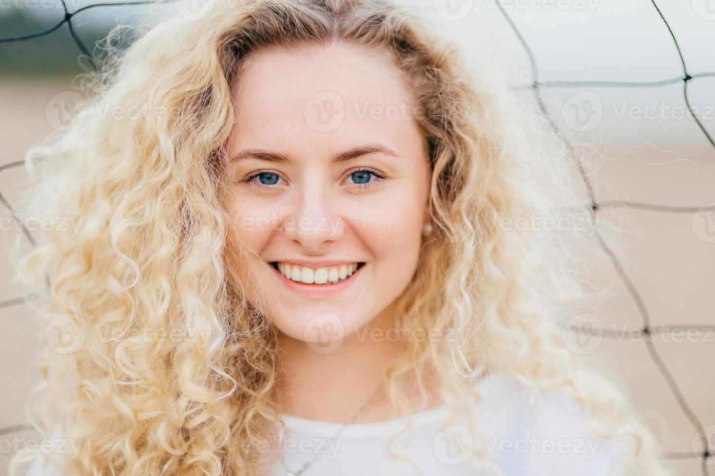 felice donna caucasica riccia con un sorriso a trentadue denti, ha capelli chiari e folti ricci, occhi azzurri, pelle pulita, sta all'aperto, ha un aspetto accattivante. primo piano ritratto di bella femmina riposa in spiaggia foto