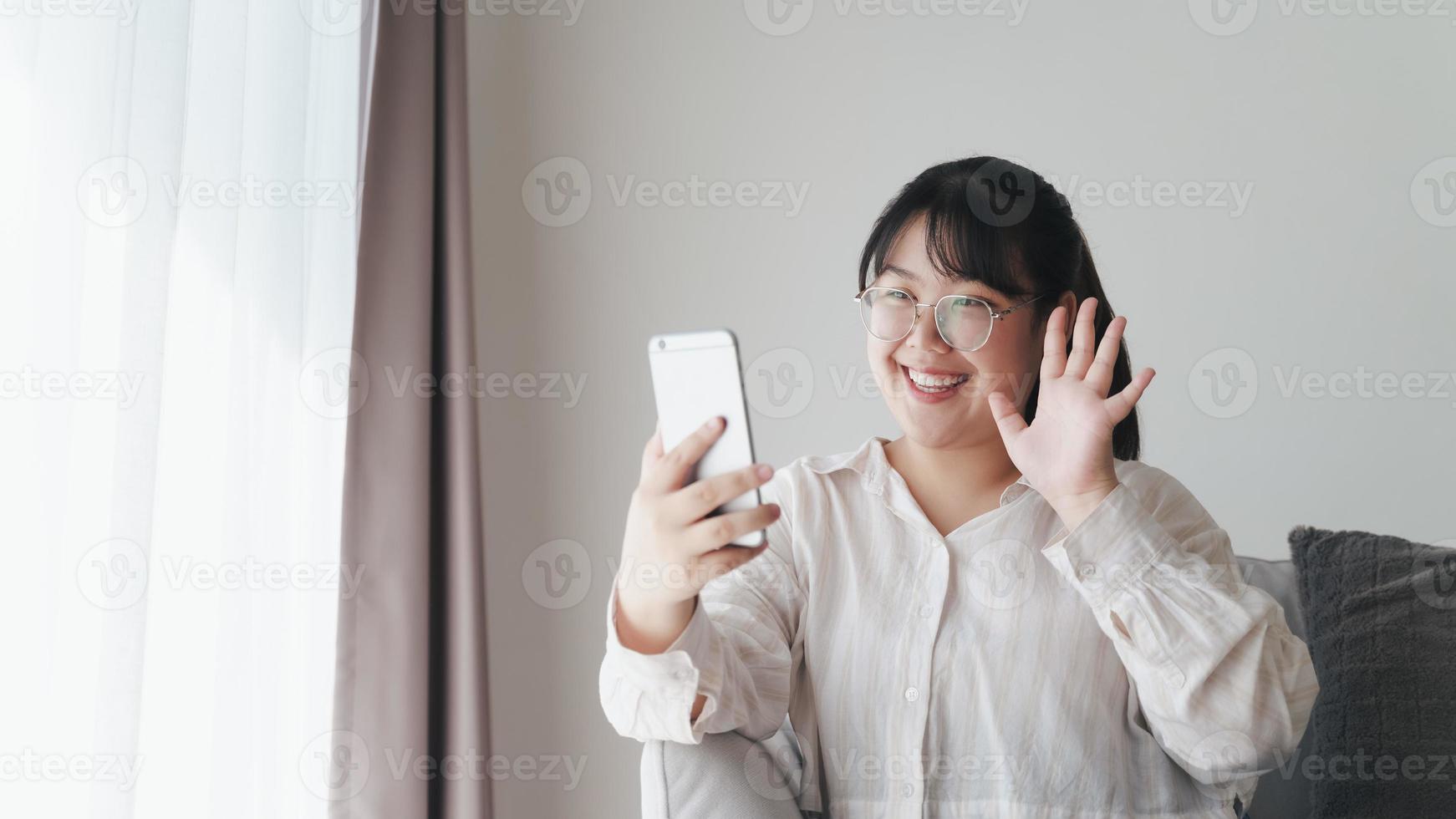 giovane donna asiatica che utilizza lo smartphone per la videoconferenza online agitando la mano facendo un gesto di saluto sul divano in soggiorno. foto